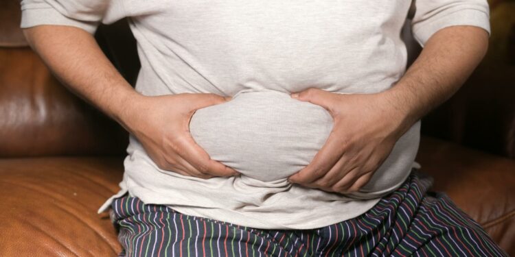A close-up view of a man holding his tummy while sitting on a couch, highlighting lifestyle choices.
