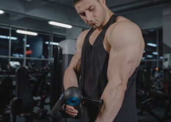 A muscular man uses a massage gun in a gym, showcasing fitness and recovery.