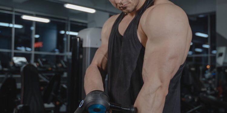 A muscular man uses a massage gun in a gym, showcasing fitness and recovery.